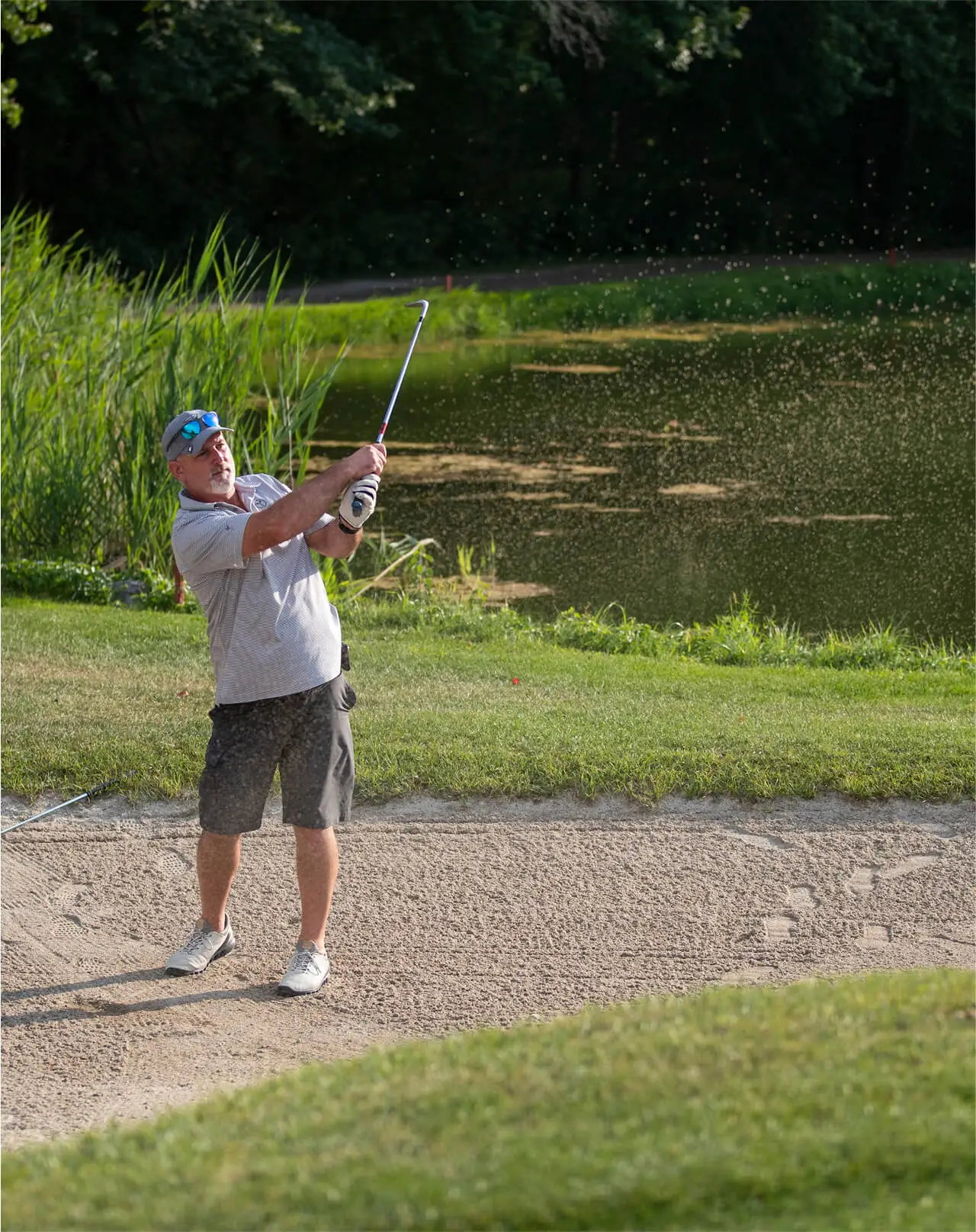 An elderly man, viewed from the front, preparing to hit a golf ball at Great Gorge Golf Club. A peaceful lake and forest are visible in the background.