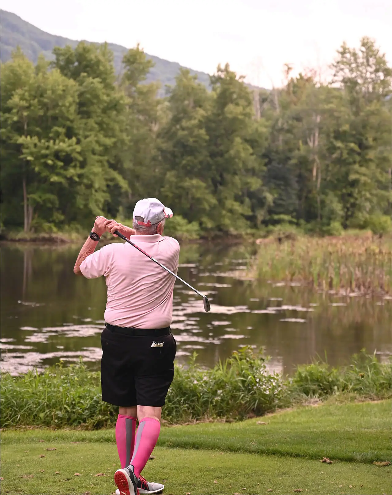 An elderly man, seen from behind, preparing to hit a golf ball at Great Gorge Golf Club. In the distance, a tranquil lake and a lush forest create a peaceful backdrop.