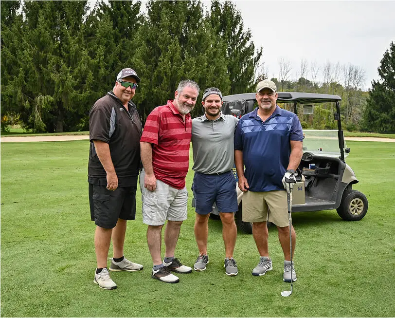 Four men posing together with a golf cart behind them at The Great Gorge in Vernon, NJ, smiling and enjoying their time on the course, with scenic fairways in the background, as. a concept of golf outings.